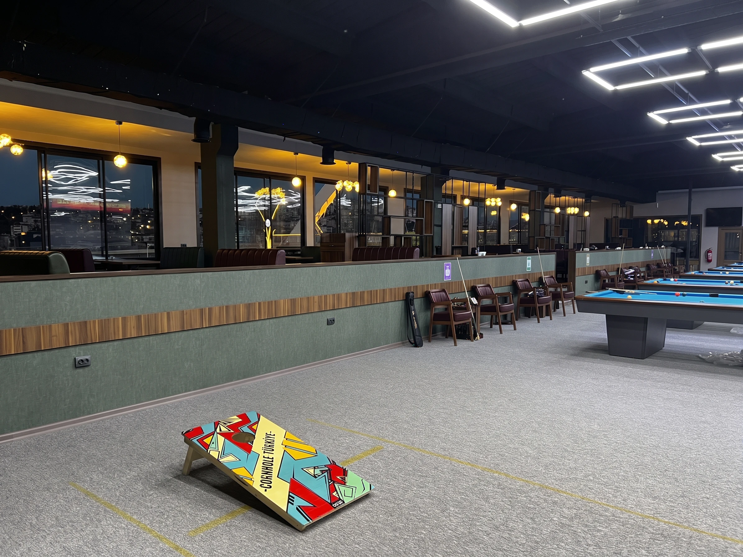 Pool salonu ve kademeli kafe platformu — cornhole sahası ve gece cephe manzarası